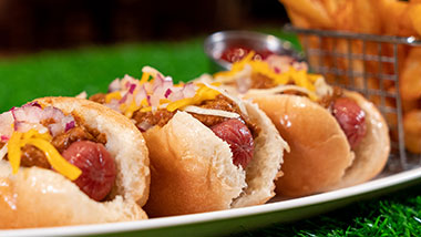 three hot dogs on a plate topped with cheese, chili and onions with fries in a basket in the background