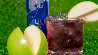 purple cocktail with apple slices next to it and a can of Red Bull in the background